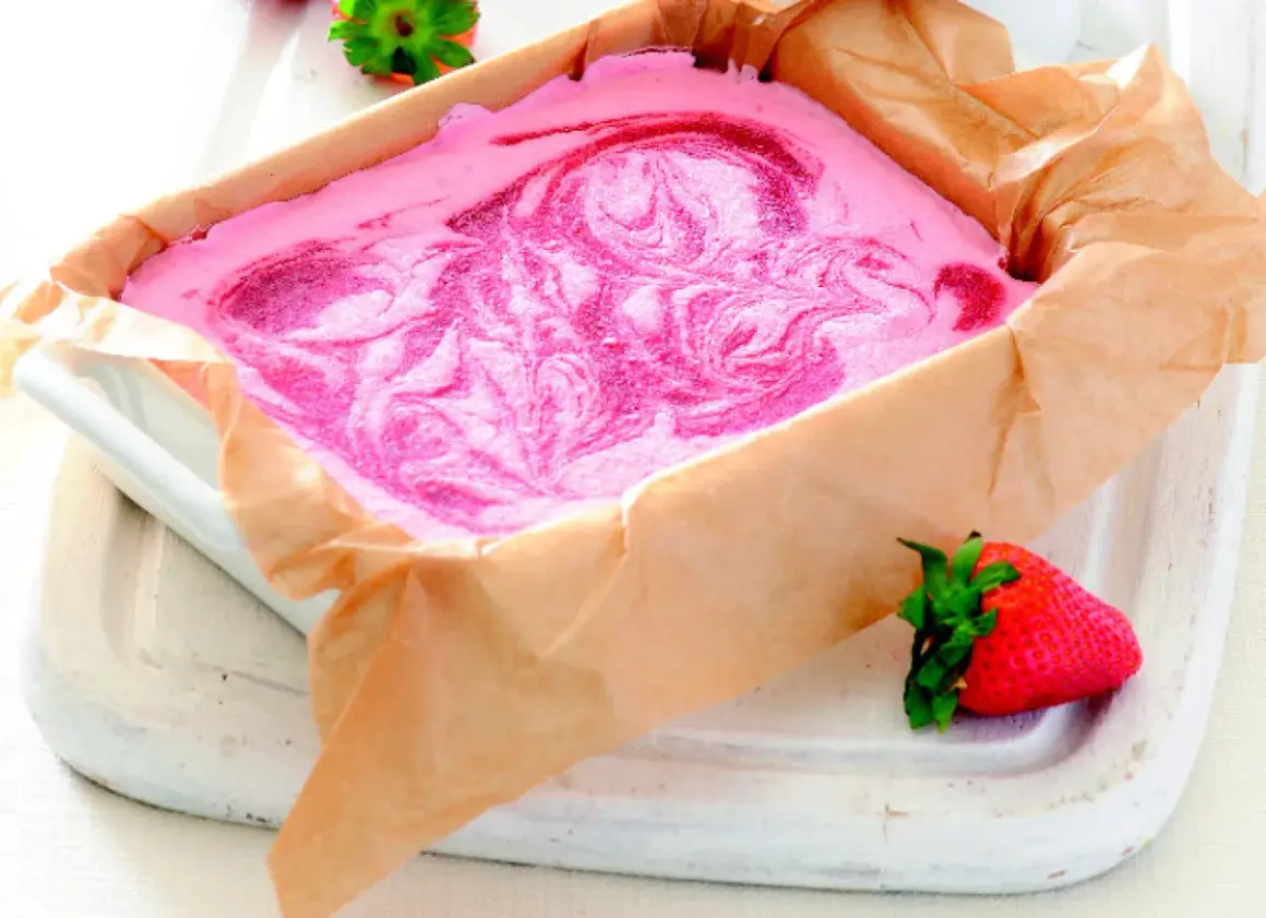 Różowe lody z mascarpone z wirami truskawkowymi w prostokątnym naczyniu wyłożonym papierem.