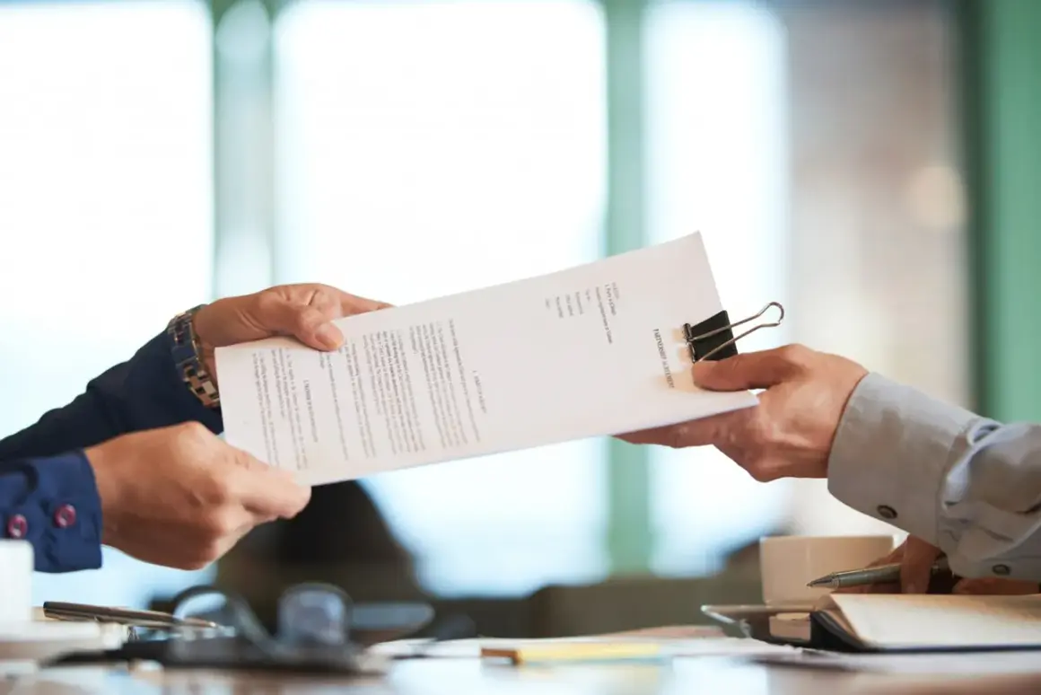 Deux mains échangent un document intitulé "PARTNERSHIP AGREEMENT", peut-être un avis d'échéance assurance, dans un bureau.