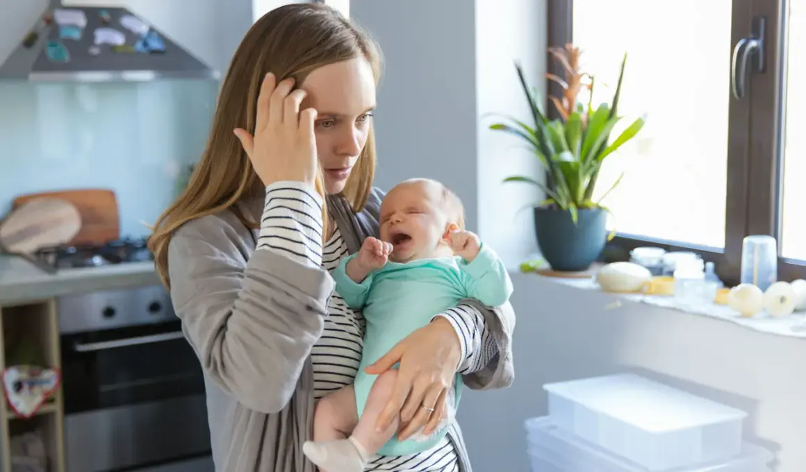 Erschöpfte Mutter hält weinendes Baby. Der 12 wochen schub macht ihr zu schaffen. Sie hält sich die Stirn.