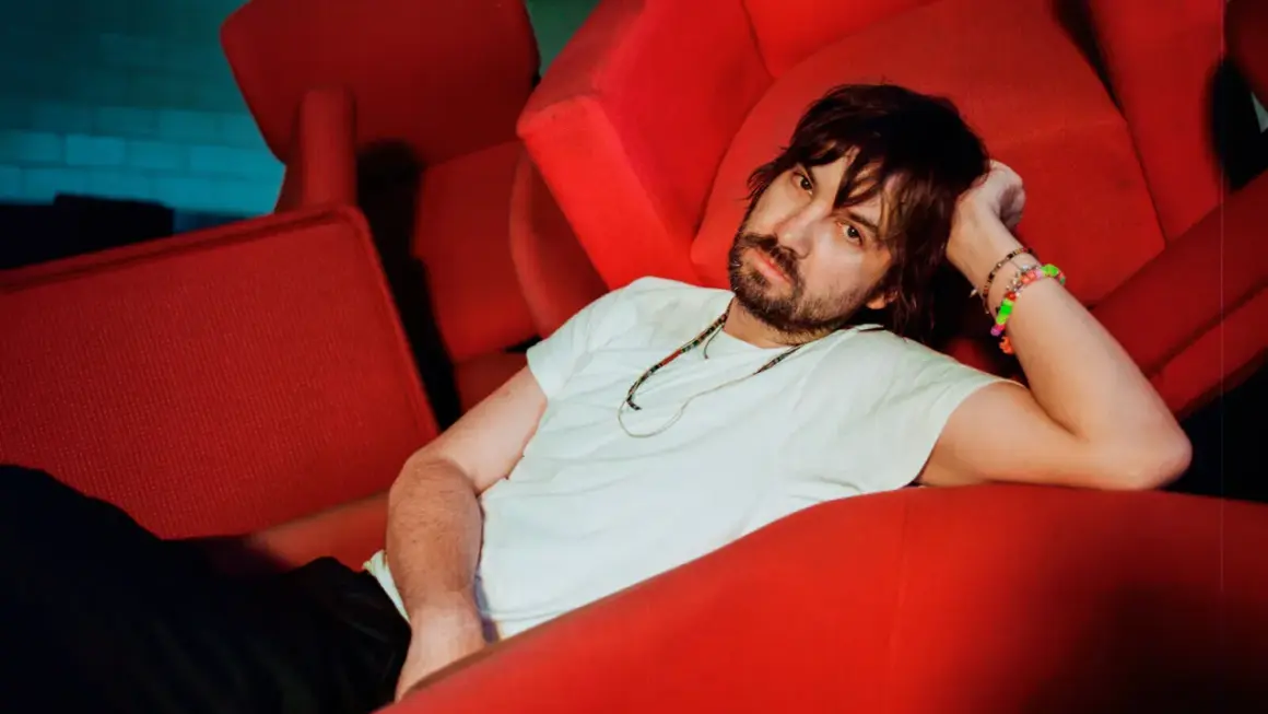 Kevin Parker z Tame Impala Polska odpoczywa na czerwonej kanapie, otoczony neonowym światłem.