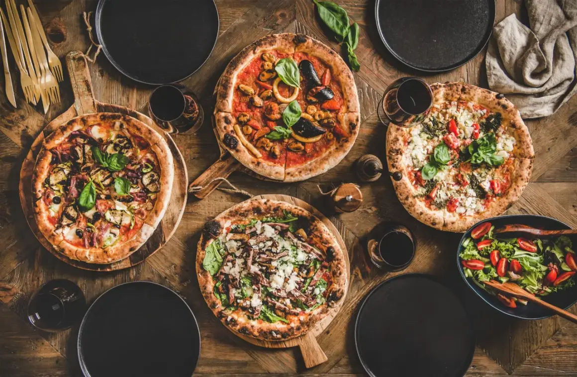 Vier Pizzen, ein Salat und Rotwein auf Holztisch. Die Frage welcher Wein zu Pizza wird hier beantwortet.