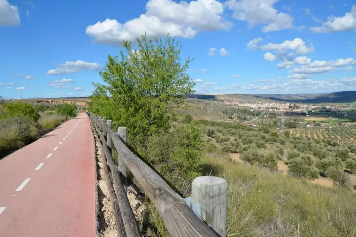 Un camino ciclista y peatonal, la **vía verde del Tajuña**, serpentea entre vegetación y un paisaje de olivos con un pueblo al fondo.