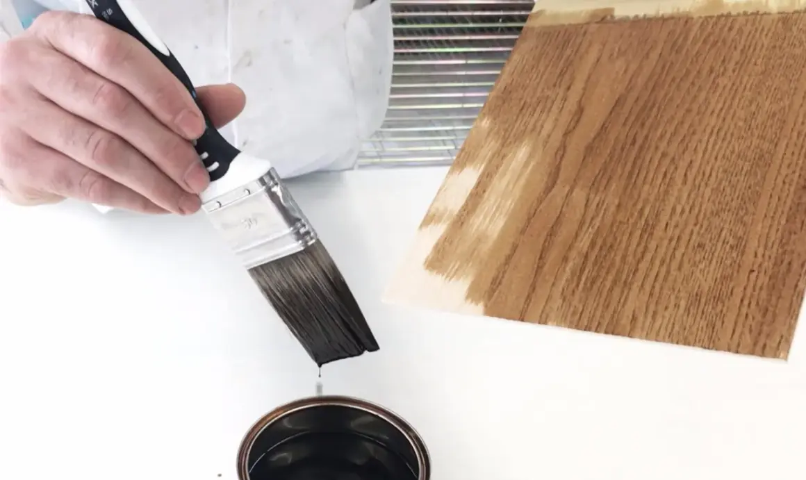 Pennello intinto in una latta di vernice scura, pronto per essere applicato su un campione di legno. Dopo l'impregnante, si mette la finitura.