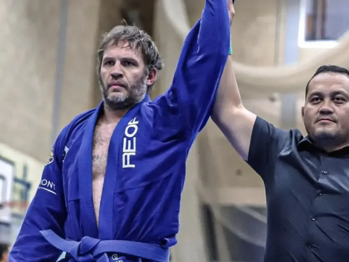Tom Hardy i blå gi, med armen lyft som segrare i BJJ.