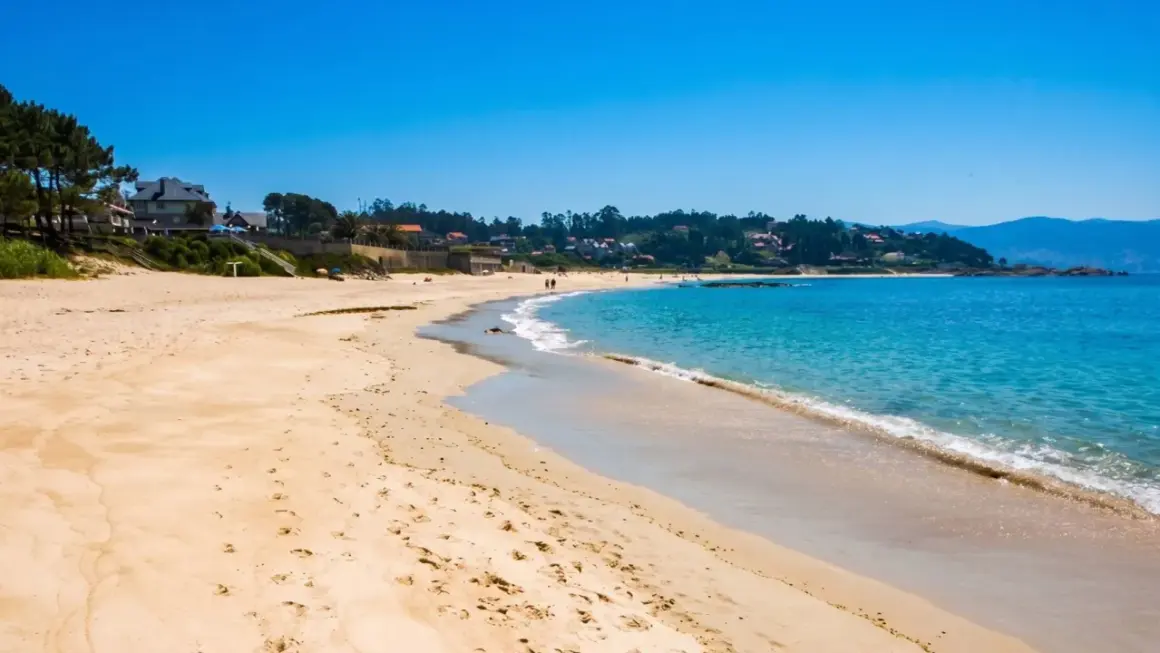 La **playa pintens** se extiende bajo un cielo azul intenso, con aguas turquesas acariciando la arena dorada. Casas y vegetación salpican la costa.