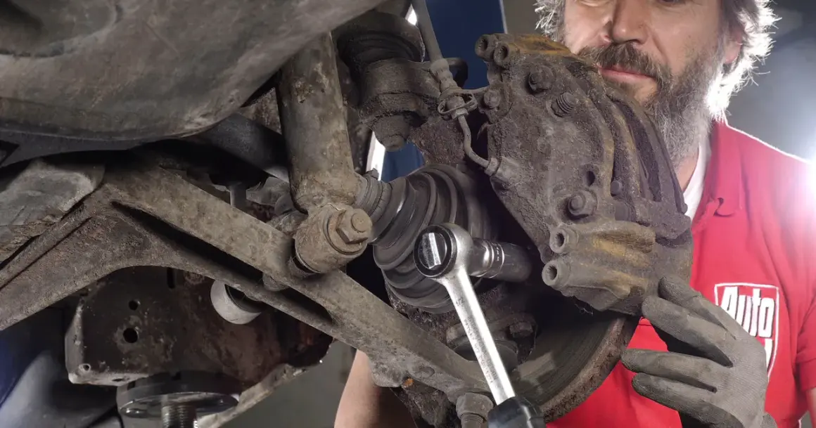 Mechanik w czerwonej koszulce z logo "Auto" dokonuje czyszczenia zacisków hamulcowych, używając klucza nasadowego.