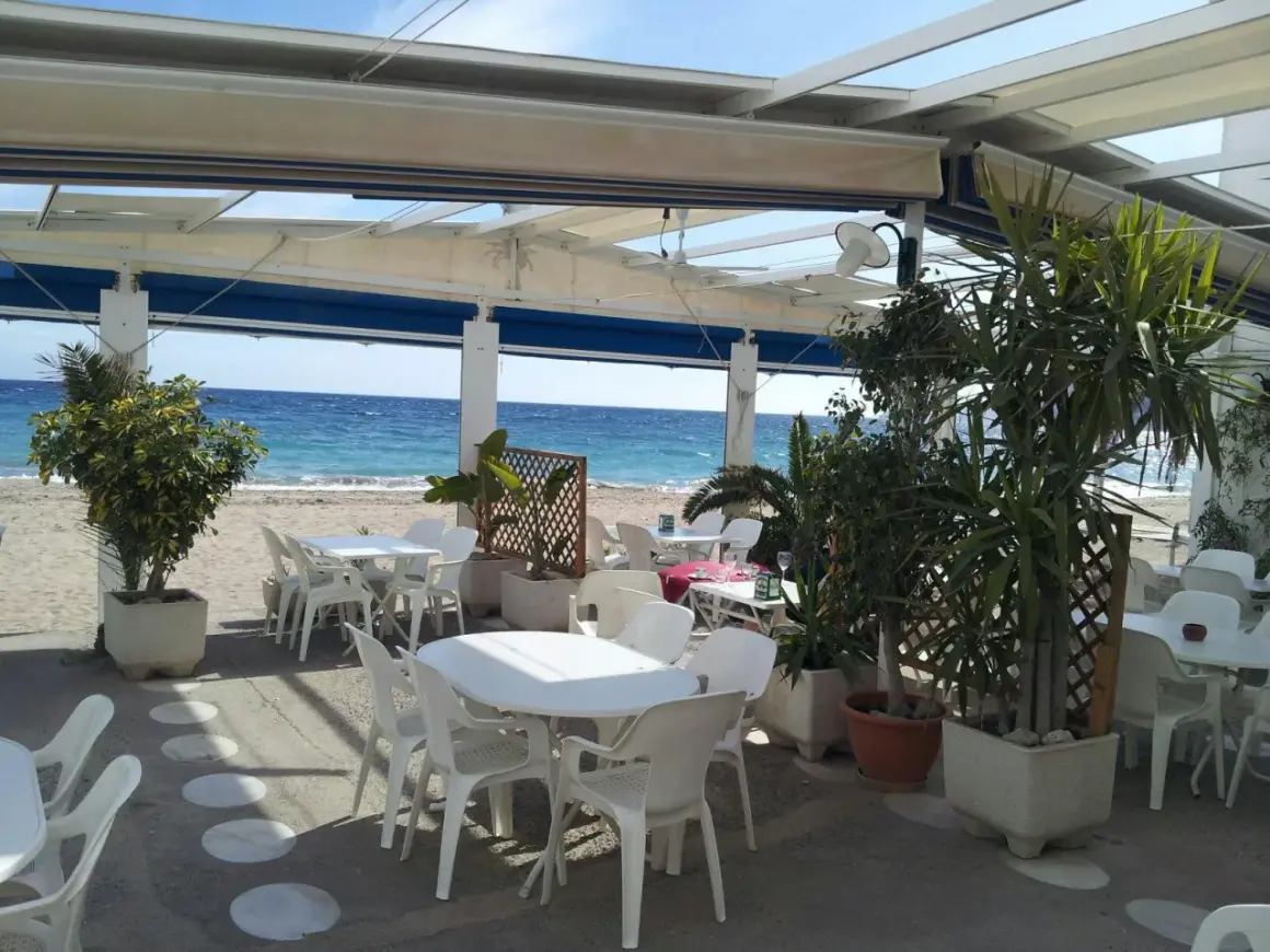 Terraza de restaurante en la playa. Mesas y sillas blancas bajo un toldo, con el mar azul de fondo. Un lugar perfecto para comer con agua amarga.