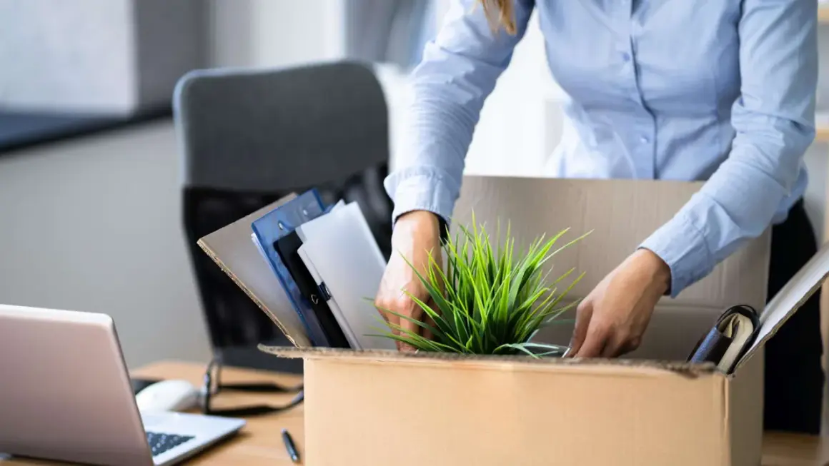 Frau packt ihre Bürosachen in einen Karton, um einen ausstand zu geben. Ein Laptop und Ordner sind zu sehen.