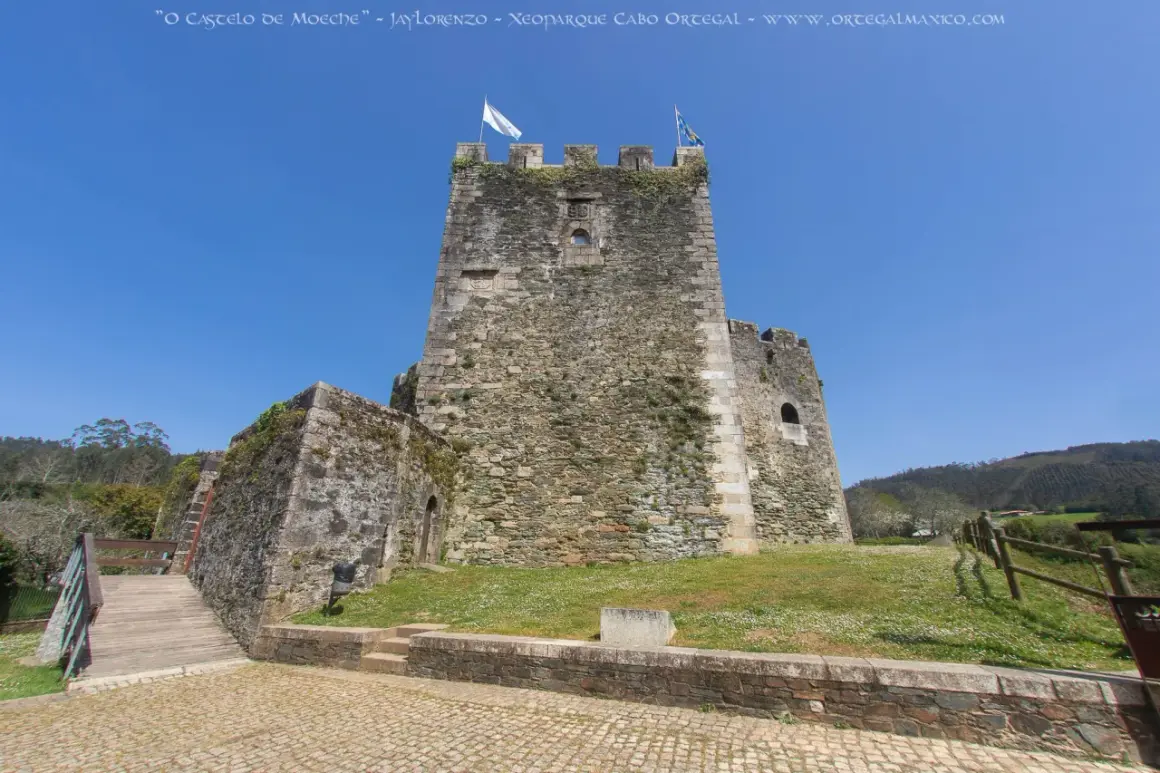 El castillo de Moeche, una imponente fortaleza de piedra, se alza bajo un cielo azul. Un puente de madera conduce a sus muros cubiertos de vegetación.
