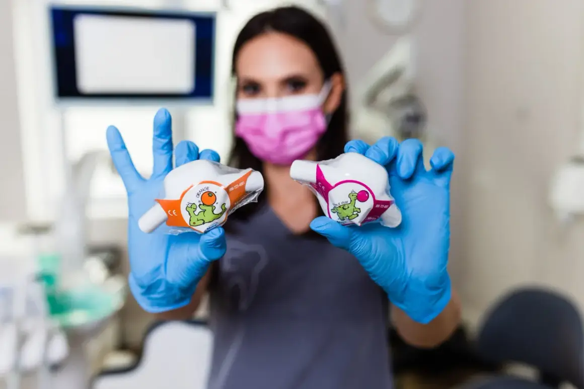 Dentystka w niebieskich rękawiczkach prezentuje dwa opakowania z zabawnymi dinozaurami, sugerujące zastosowanie w leczeniu, być może z użyciem podtlenku azotu.