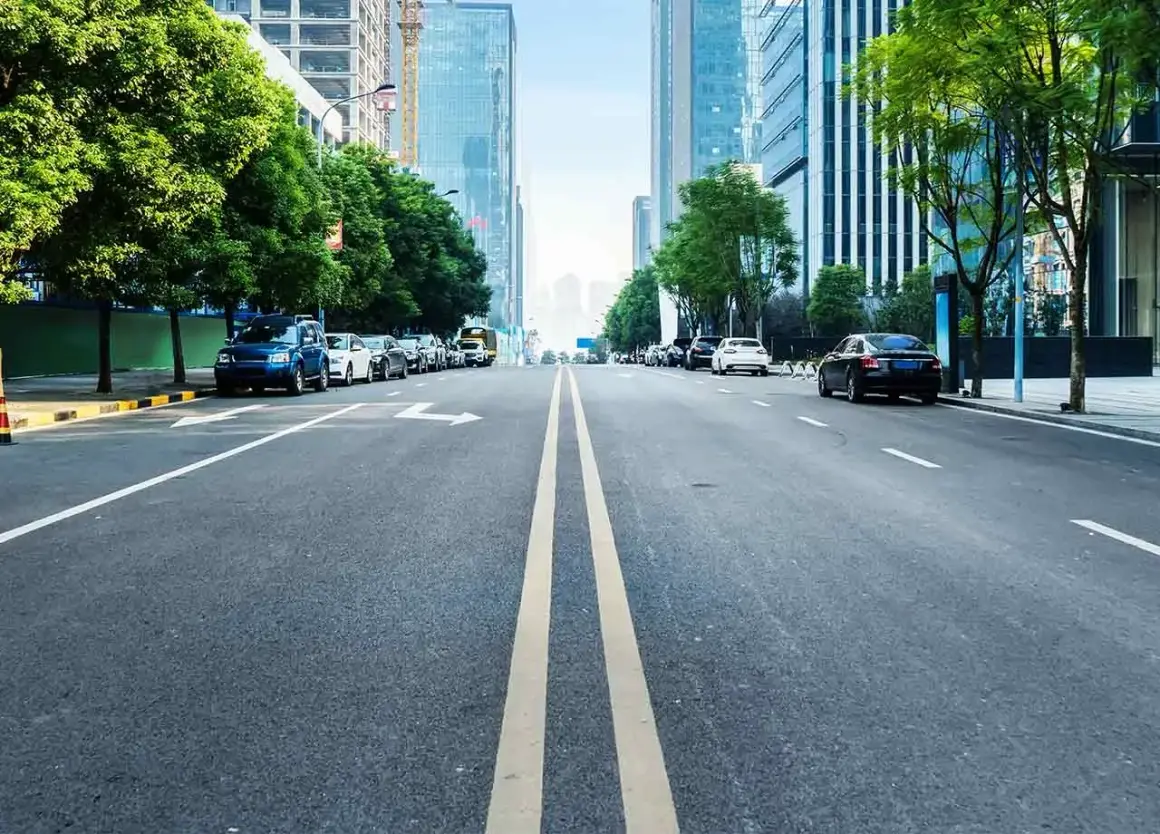 Eine Straße mit einer **durchgezogenen Linie** in der Mitte, gesäumt von Bäumen und modernen Gebäuden. Autos parken am Straßenrand.