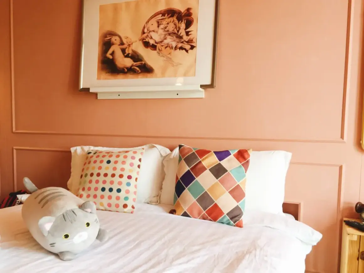 Chambre couleur terracotta avec un lit douillet, des coussins colorés et une peluche chat. Un tableau de lapins orne le mur.