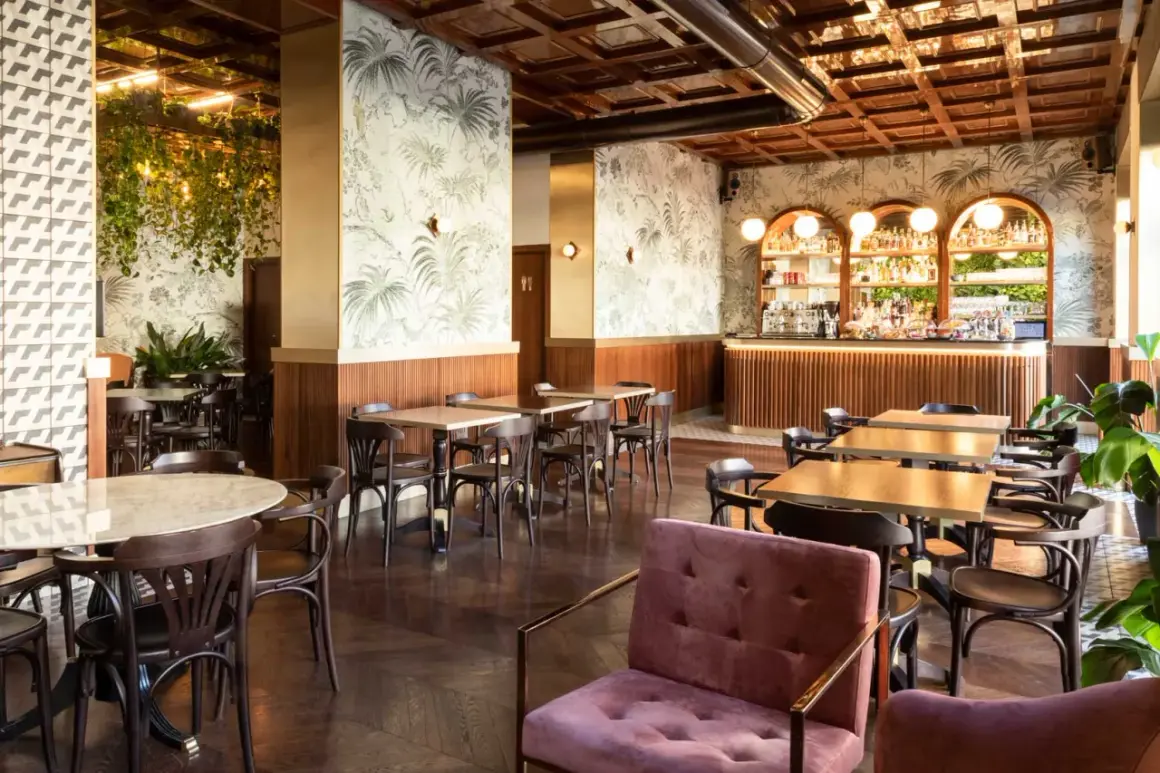 Interno di un locale con carta da parati tropicale, bancone bar illuminato e tavoli in legno. Perfetto per scoprire i **cheap bars in Milan**.