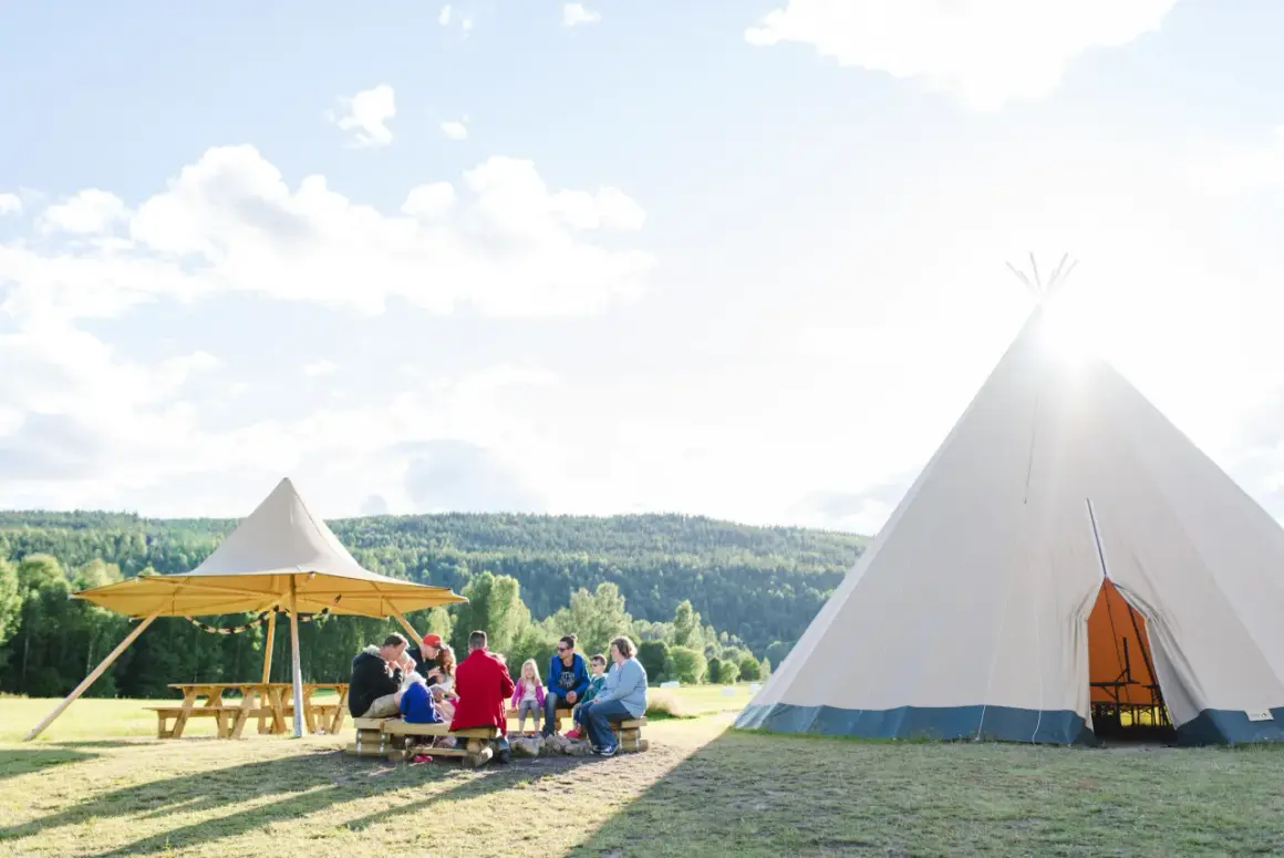 En familj njuter av en solig dag på alevi camping i Sverige. De sitter runt en lägereld under ett stort parasoll, med en tipi i bakgrunden.