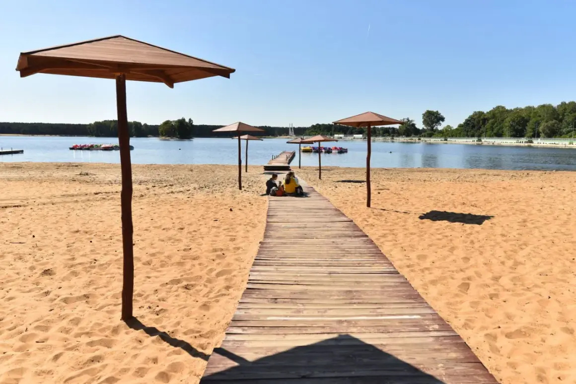 Sielpia jezioro: drewniana ścieżka prowadzi na piaszczystą plażę pod parasolami. W oddali widać pomost z rowerami wodnymi.