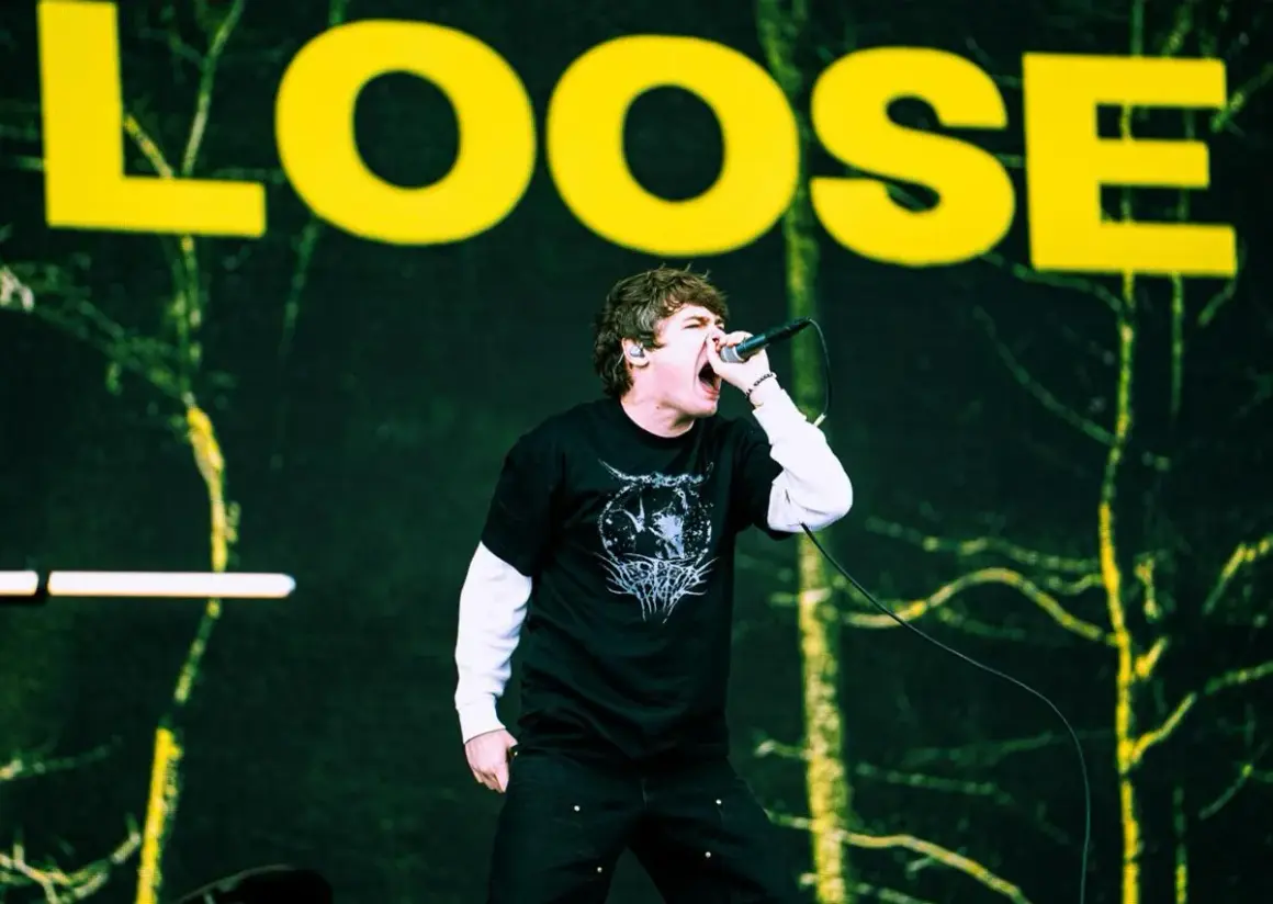 Wokalista z otwartymi ustami śpiewa do mikrofonu na tle sceny z napisem "LOOSE".