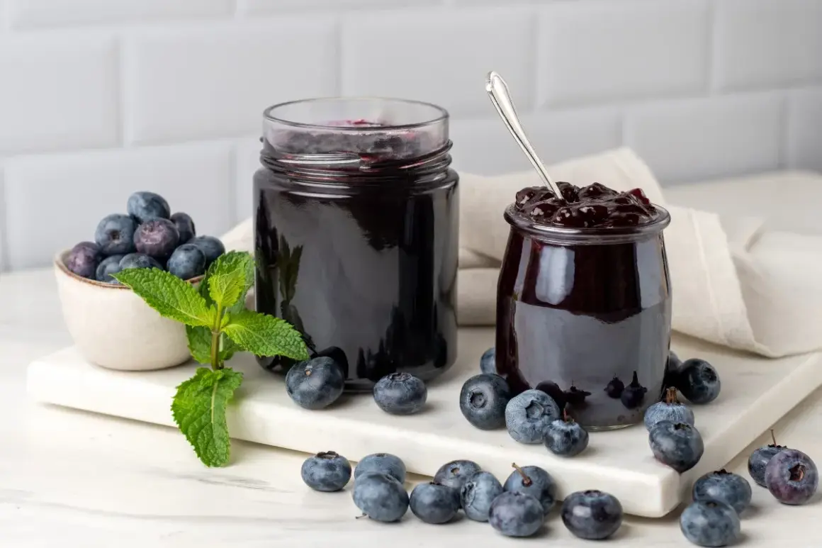 Due barattoli di marmellata di mirtilli, uno aperto con un cucchiaino, circondati da mirtilli freschi e foglie di menta.