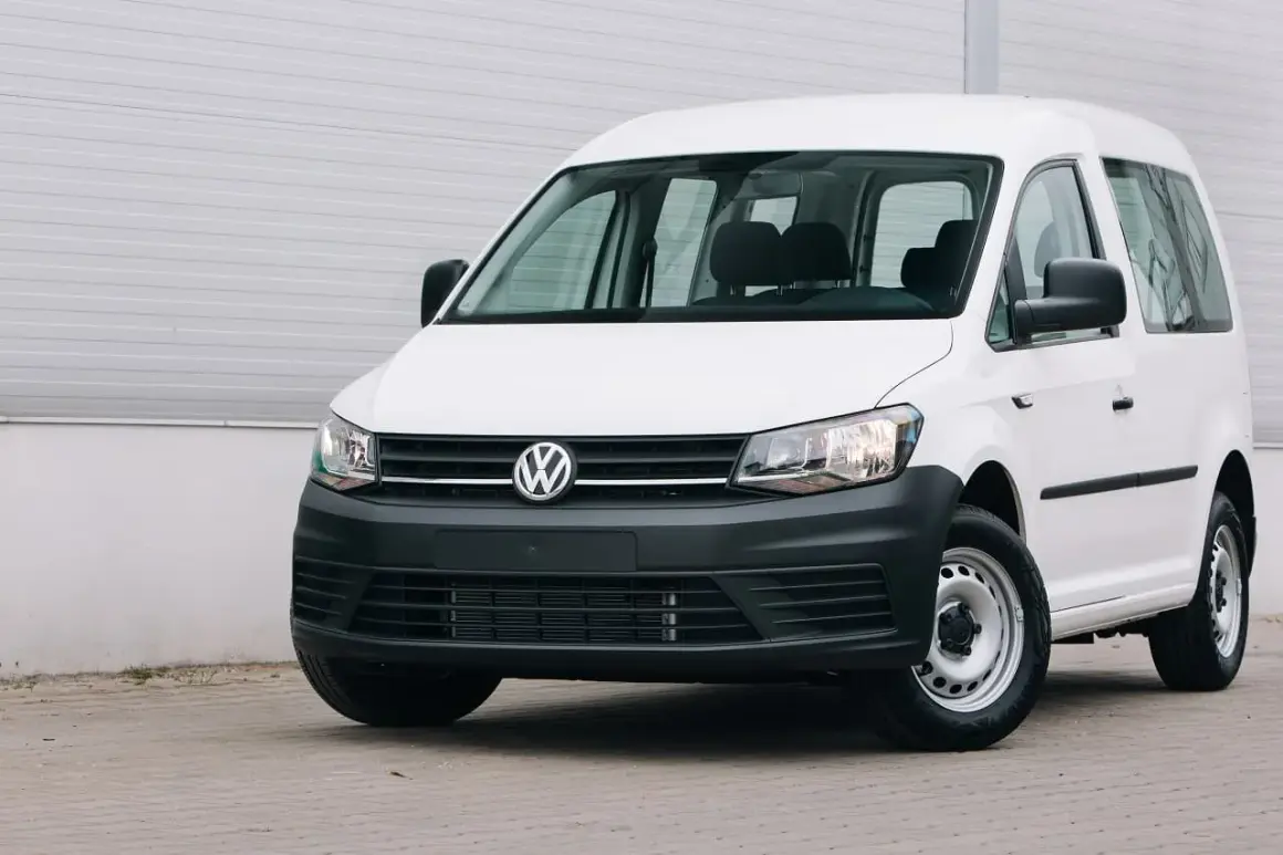Biały Volkswagen Caddy, samochód osobowy zarejestrowany jako ciężarowy bez kratki, stoi przed szarą ścianą.