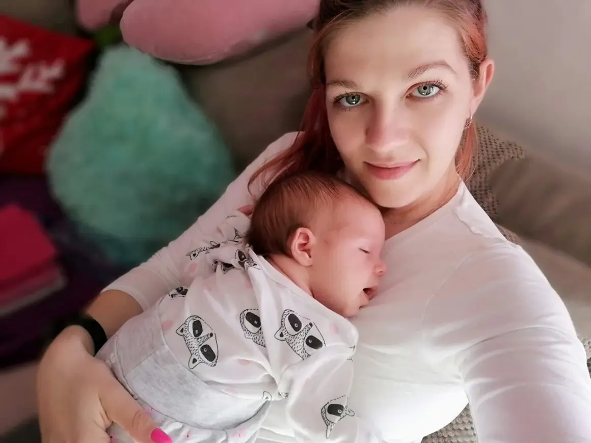 Mama tuli otula swoje śpiące dziecko, czułe kangurowanie dziecka w białym pajacyku z motywem zwierzątek.