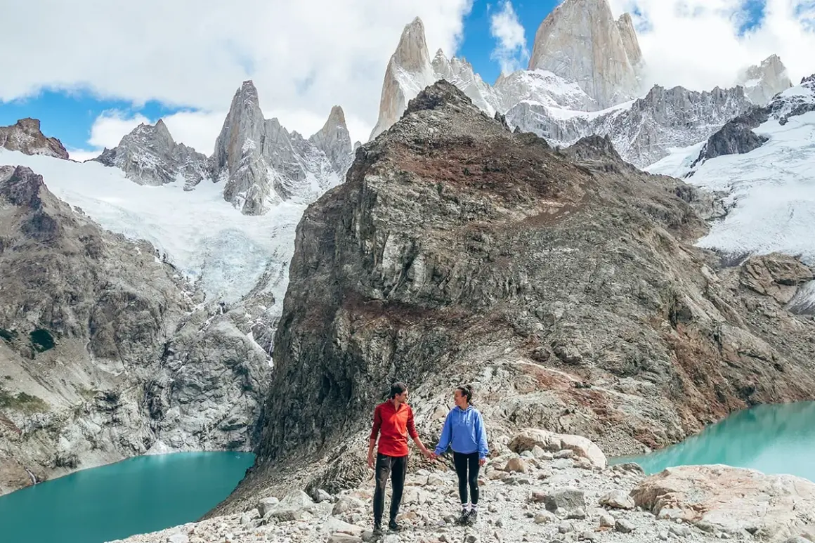 Deux randonneurs se tiennent la main sur un sentier rocheux, admirant un lac turquoise et des montagnes enneigées lors d'une randonnée argentine.