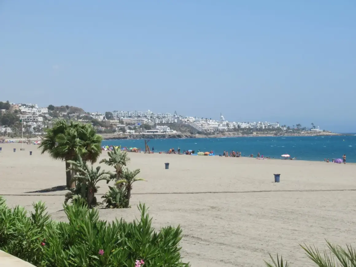 La **playa de la Mena** se extiende con arena dorada y palmeras. Al fondo, un pueblo blanco se asoma al mar azul.