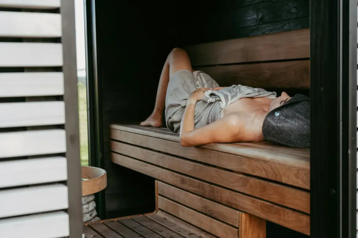 Kobieta w czepku "OF SAUNA" leży na drewnianej ławie w saunie, relaksując się. Dowiedz się, jak oddychać w saunie, by w pełni czerpać z jej dobrodziejstw.