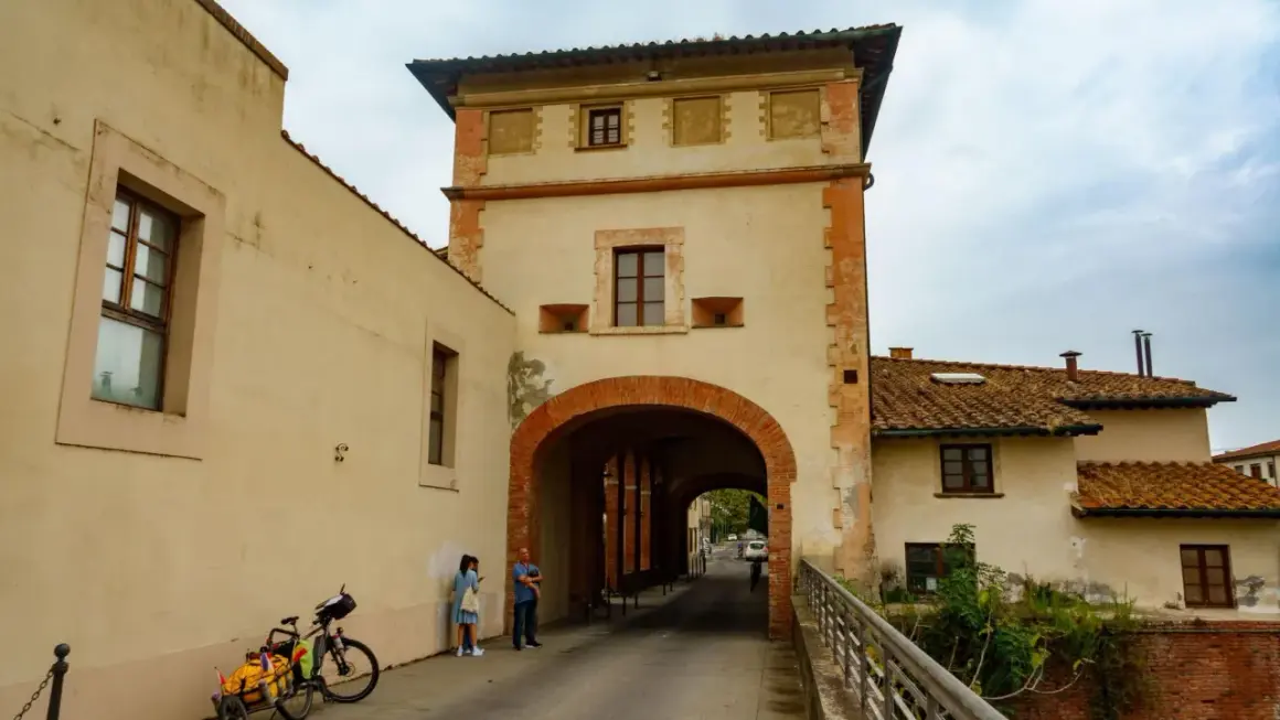 Pieve a Nievole: un arco in mattoni rossi conduce a una strada con case antiche. Una coppia e una bici con carretto attendono.