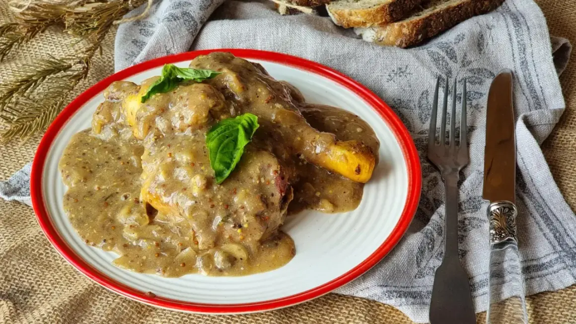 Delicioso pollo a la mostaza cubierto con una cremosa salsa, servido en un plato blanco con borde rojo. Acompañado de pan y cubiertos.