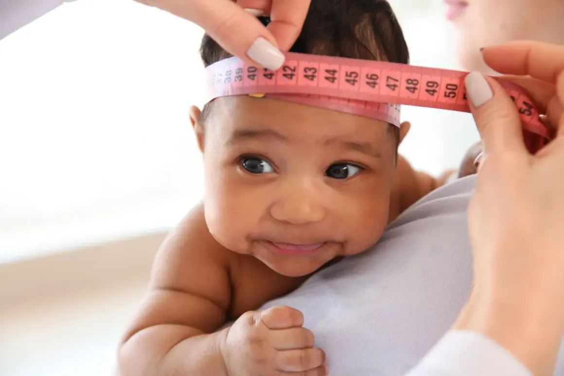 Ein Baby mit dunkler Haut wird der Kopfumfang gemessen. Ein Maßband zeigt etwa 40 cm.