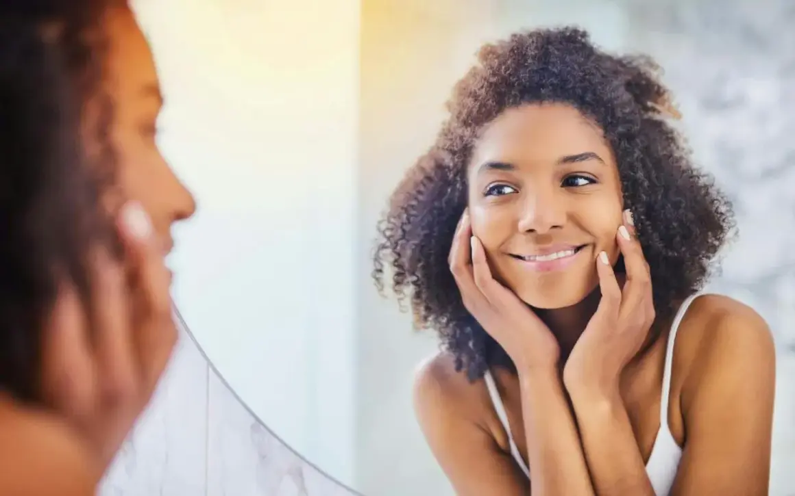 Une femme aux cheveux bouclés se regarde dans un miroir, les mains posées sur ses joues, comme pour se masser le visage. Elle sourit doucement.