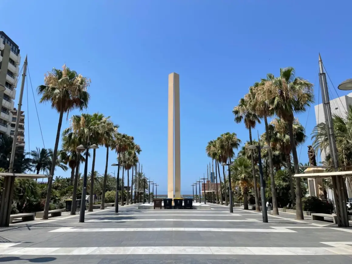 Un obelisco se alza en la plaza de las velas Almería, flanqueado por palmeras y un cielo azul despejado.