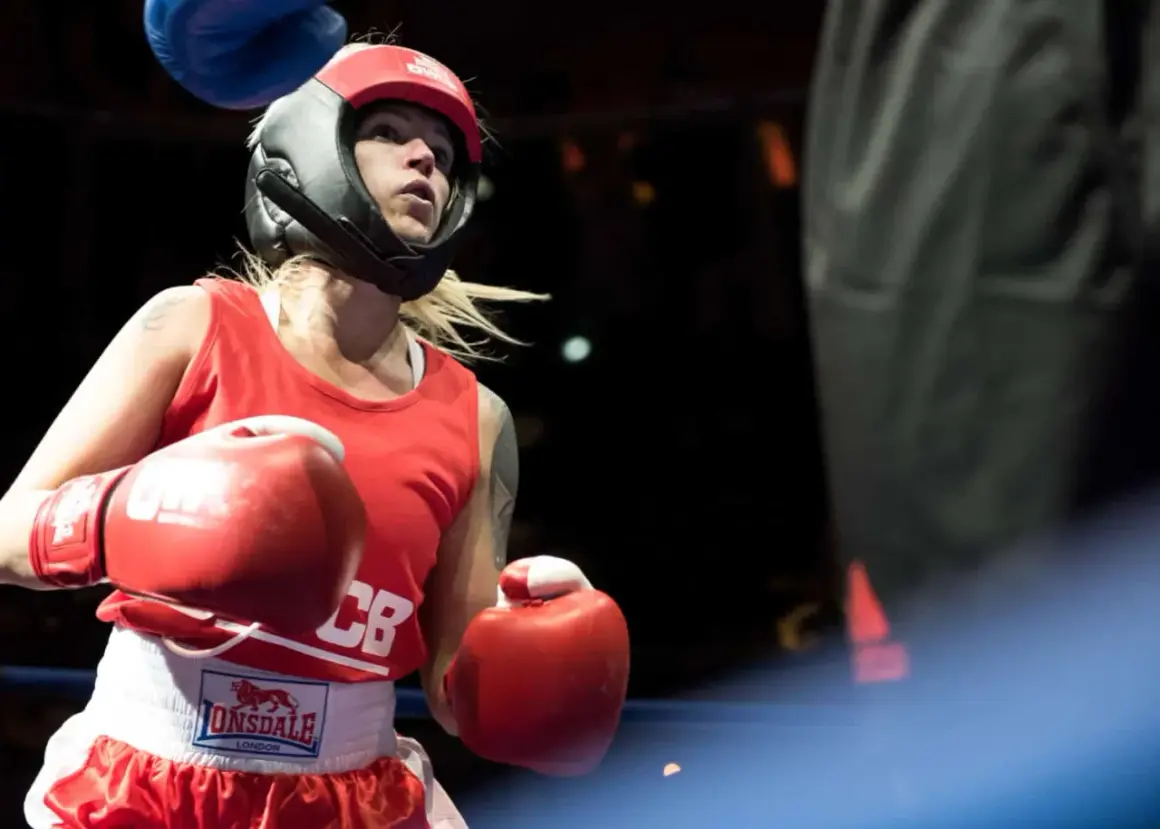 A determined female boxer in red gear and headguard prepares for a white collar boxing match, her red gloves raised.