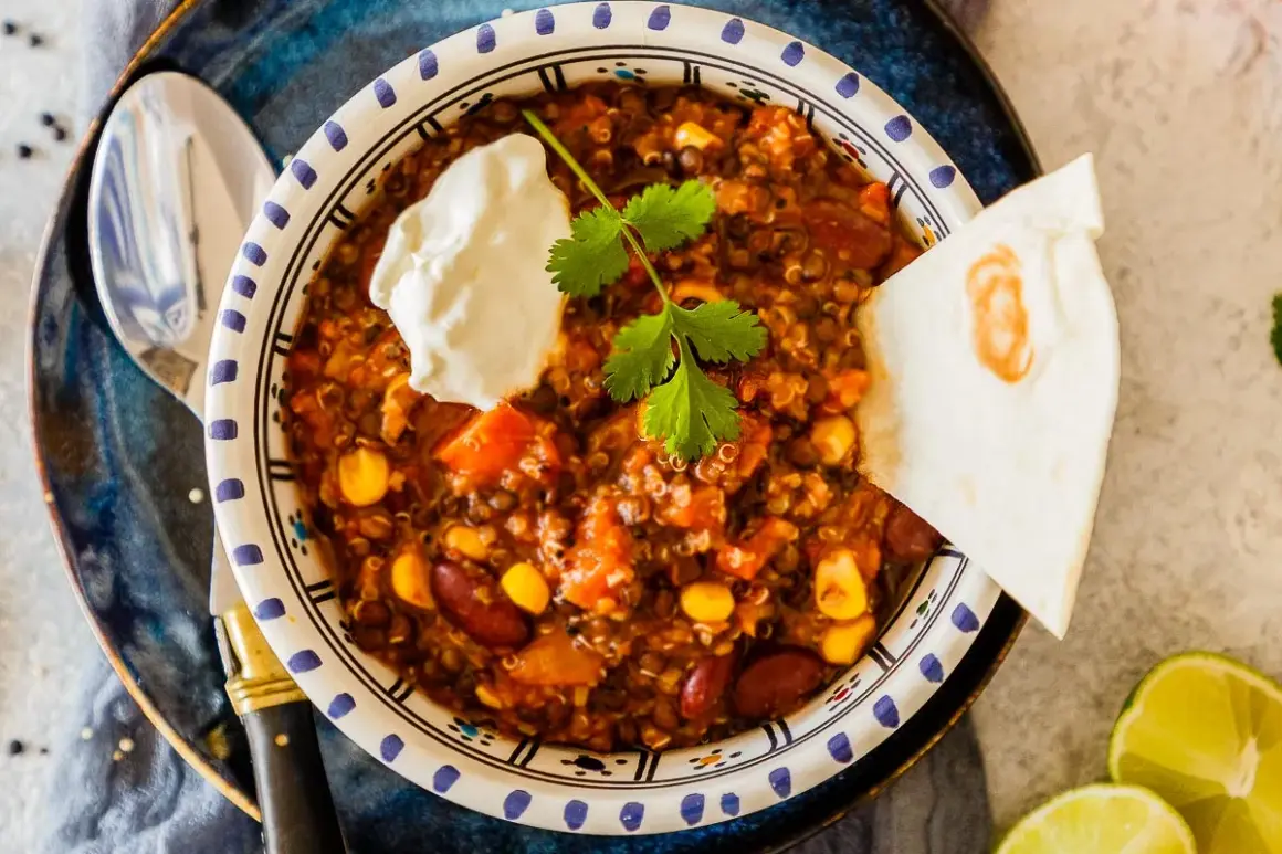 Herzhaftes Chili sin Carne mit Quinoa, Bohnen und Mais, garniert mit Sauerrahm und Koriander. Dazu eine Tortilla-Ecke und Limettenspalten.