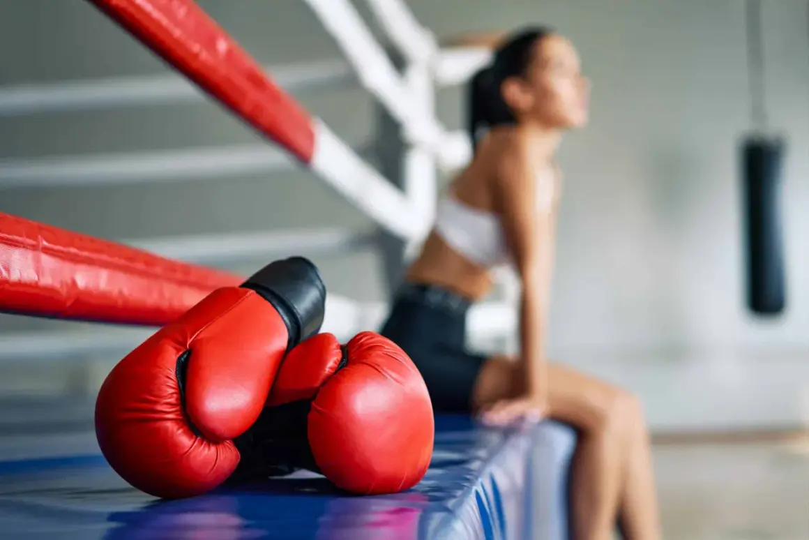 Red boxing gloves rest on the edge of a ring, hinting at a boxer puncher style. A woman sits in the background, her face turned away.