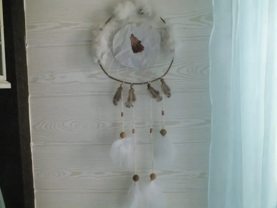 Un attrape rêve, ornement traditionnel, orné d'un loup hurlant, de plumes et de perles, suspendu devant un mur en bois clair.