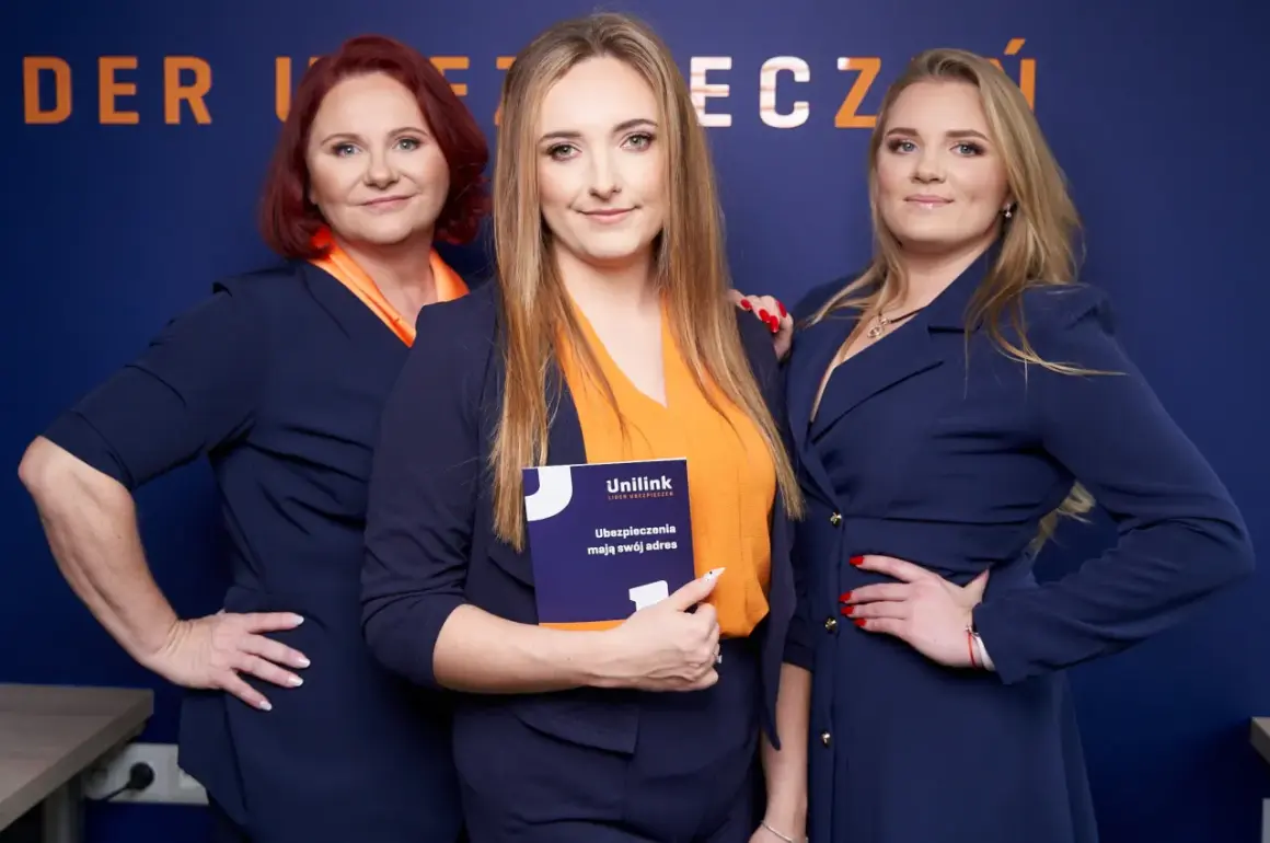 Trzy kobiety w granatowych marynarkach i pomarańczowych bluzkach pozują na tle niebieskiej ściany z napisem.