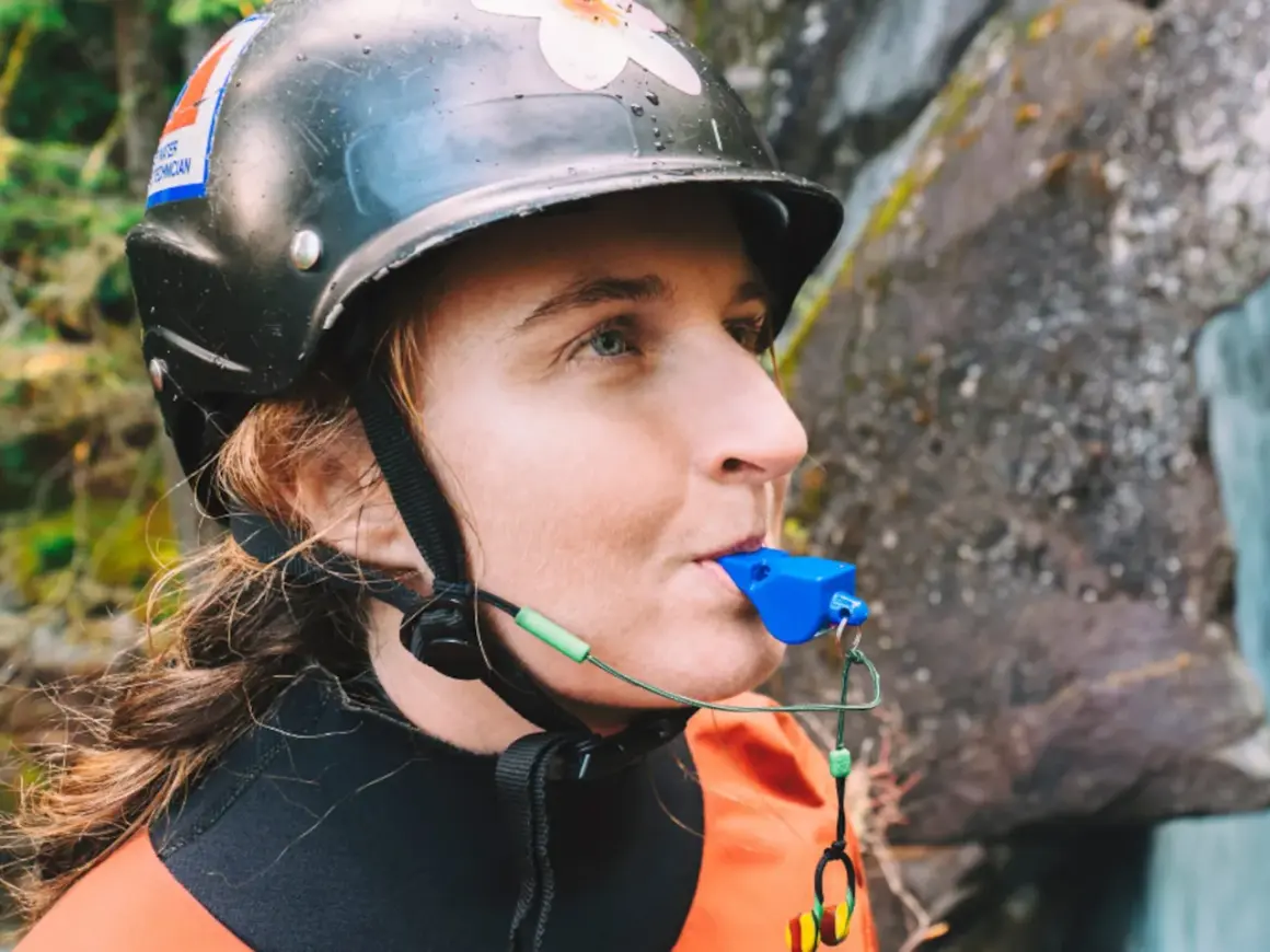Persona con casco y traje naranja sopla un silbato de emergencia azul, listo para cualquier aventura acuática.