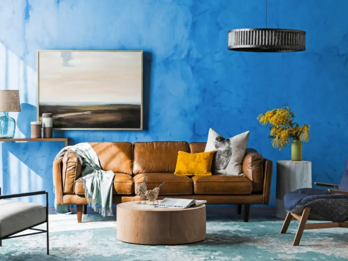 Ein Wohnzimmer mit leuchtend blauer Wand. Ein braunes Ledersofa, ein gelbes Kissen und ein Bild mit Landschaftsmotiv. Welche Farbe passt gut zu Blau? Hier: Gelb und Braun.