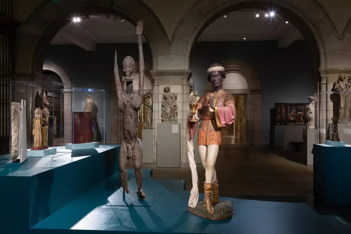 Two striking sculptures, one a wooden figure with raised arms, the other a painted figure holding a chalice, are displayed in a museum gallery, showcasing medieval African art.