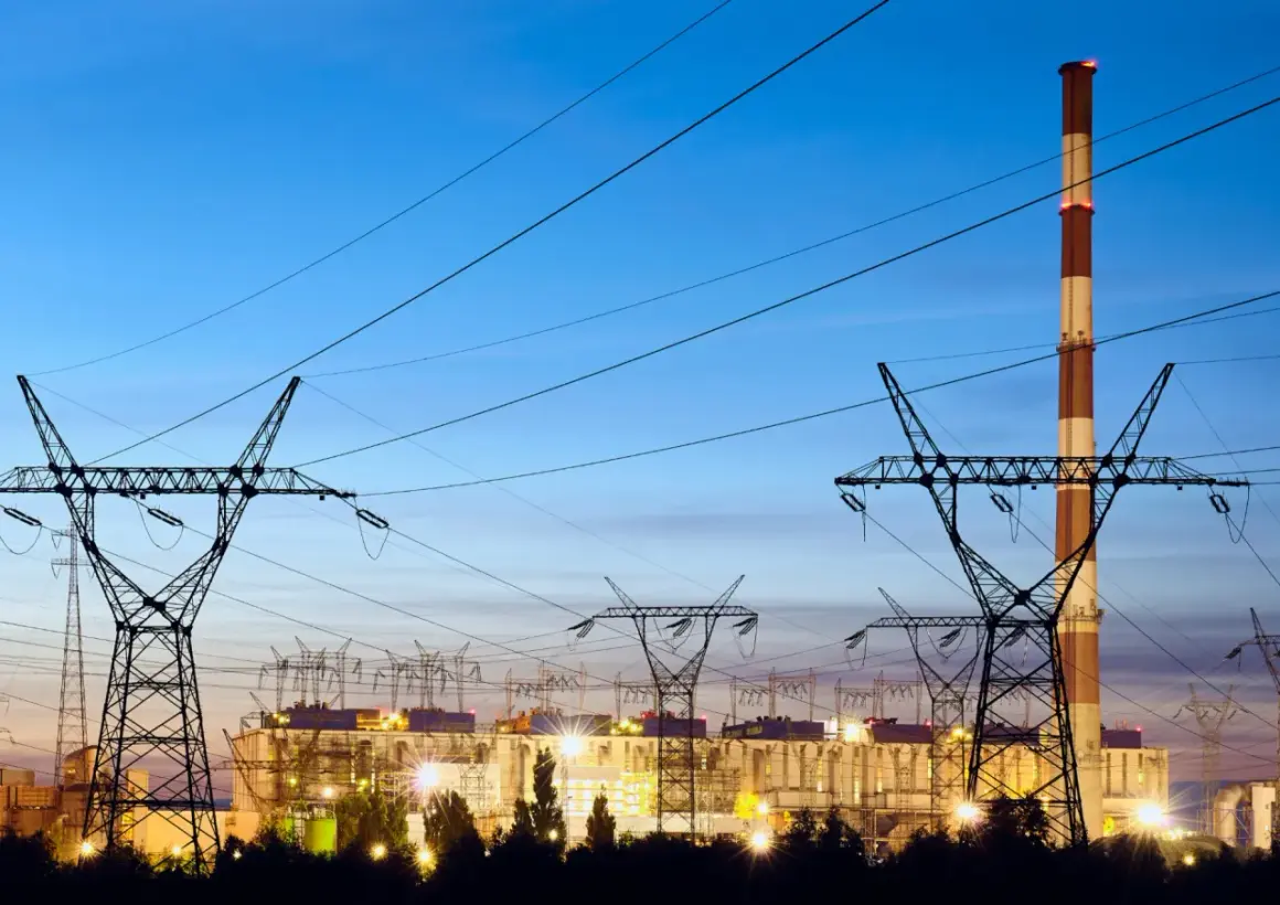 Elektrownia z kominem i liniami wysokiego napięcia. W tle widać budynki zakładu, a nad nimi niebo o zmierzchu. To obraz, który może kojarzyć się z opłatą kogeneracyjną.