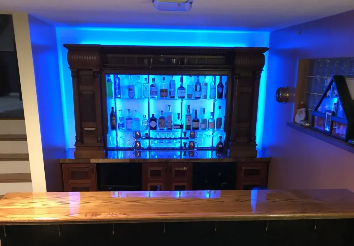 A stunning home bar setup, perfect for entertaining. This image shows how to build a bar with a wooden countertop, a backlit liquor cabinet filled with bottles, and vibrant blue ambient lighting.