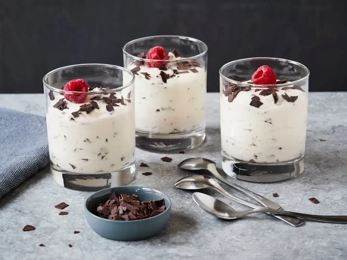 Drei Gläser mit cremigem Dessert, garniert mit Himbeeren und Schokoladenstückchen. Ein Schälchen mit Schokostückchen und Löffel daneben. Ein leckeres Herrencreme Rezept zum Nachmachen.