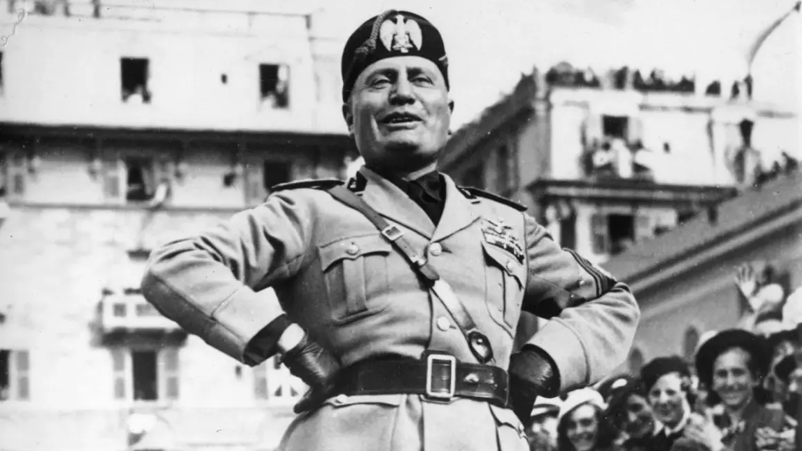 Benito Mussolini, con uniforme militar y gorra, sonríe ante una multitud. Su ascenso al poder es un capítulo oscuro de la historia.