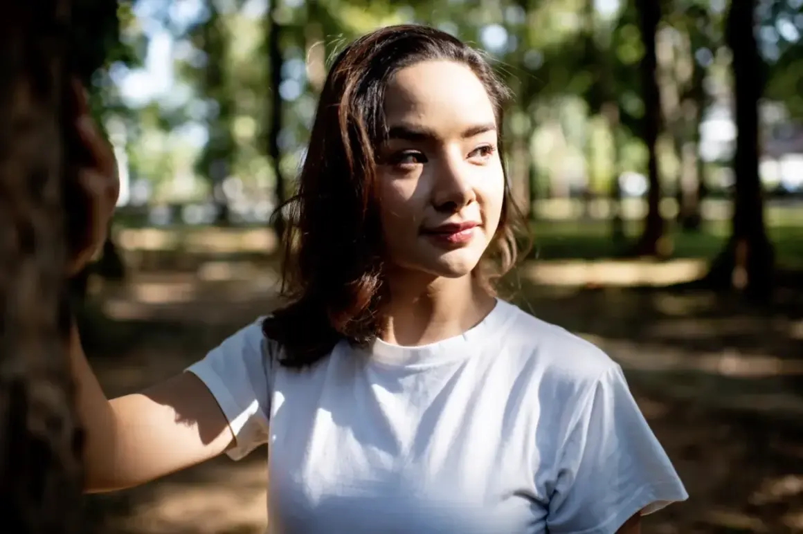 A young woman in a white t-shirt stands in a park, her face illuminated by dappled sunlight, showcasing the dramatic contrast of soft lighting vs hard lighting.