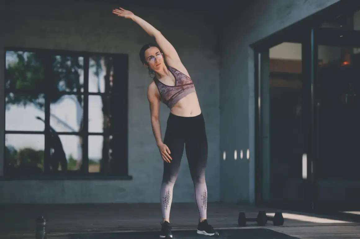 Femme faisant des étirements pour sa séance de musculation maison. Elle est en tenue de sport, bras levé, penchée sur le côté.
