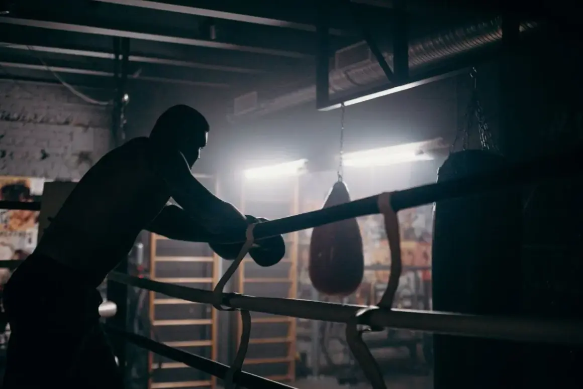 A boxer leans on the ropes in a dimly lit gym, contemplating how to get into boxing matches. Punching bags hang in the background.