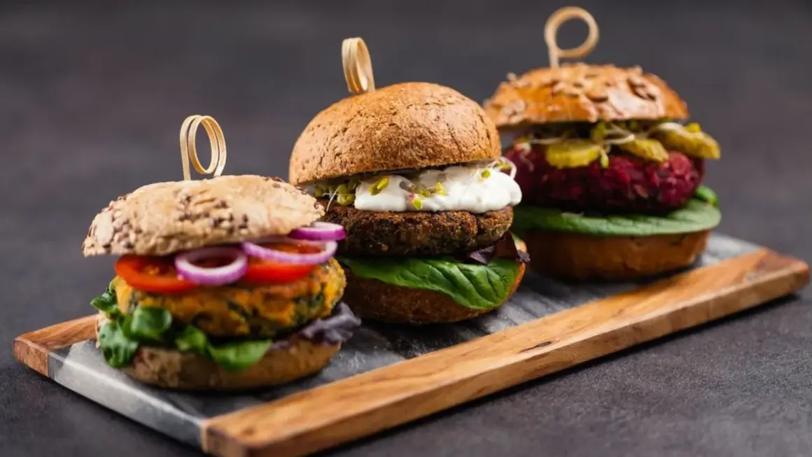 Drei appetitliche vegetarische Gerichte: Burger mit bunten Patties, frischem Gemüse und cremigen Saucen auf rustikalem Holzbrett.