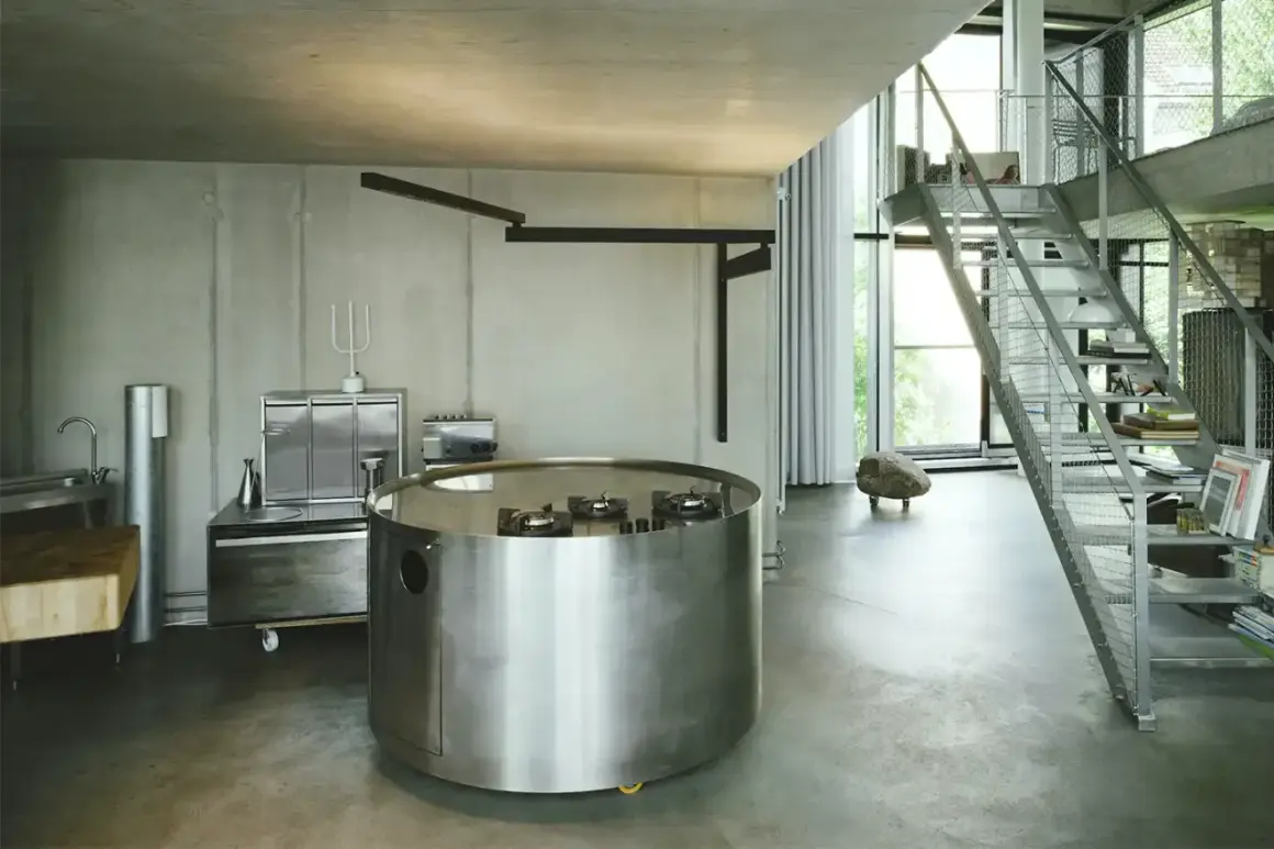 Interno in stile brutalista con cucina circolare in metallo, scale a vista e pareti in cemento grezzo.
