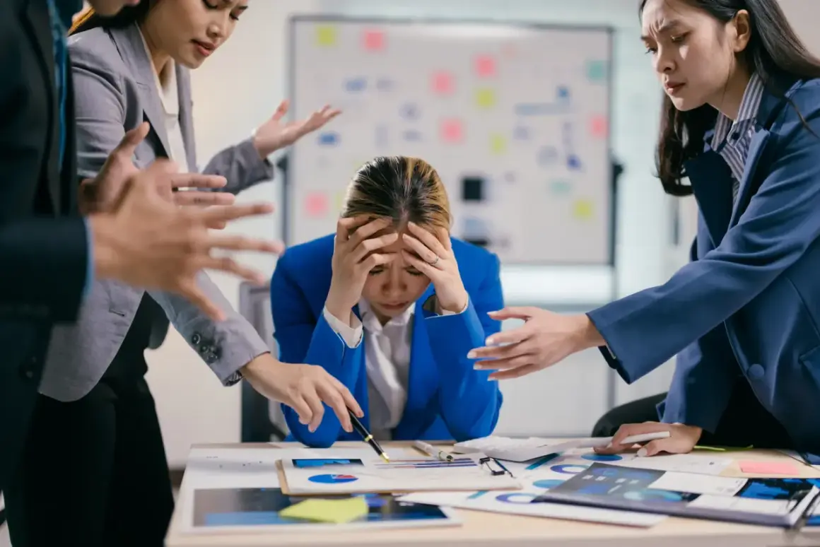 Giovane donna d'affari stressata, con le mani sulla testa, mentre colleghi la circondano con gesti agitati. Lo stress lavoro correlato è un rischio da valutare.