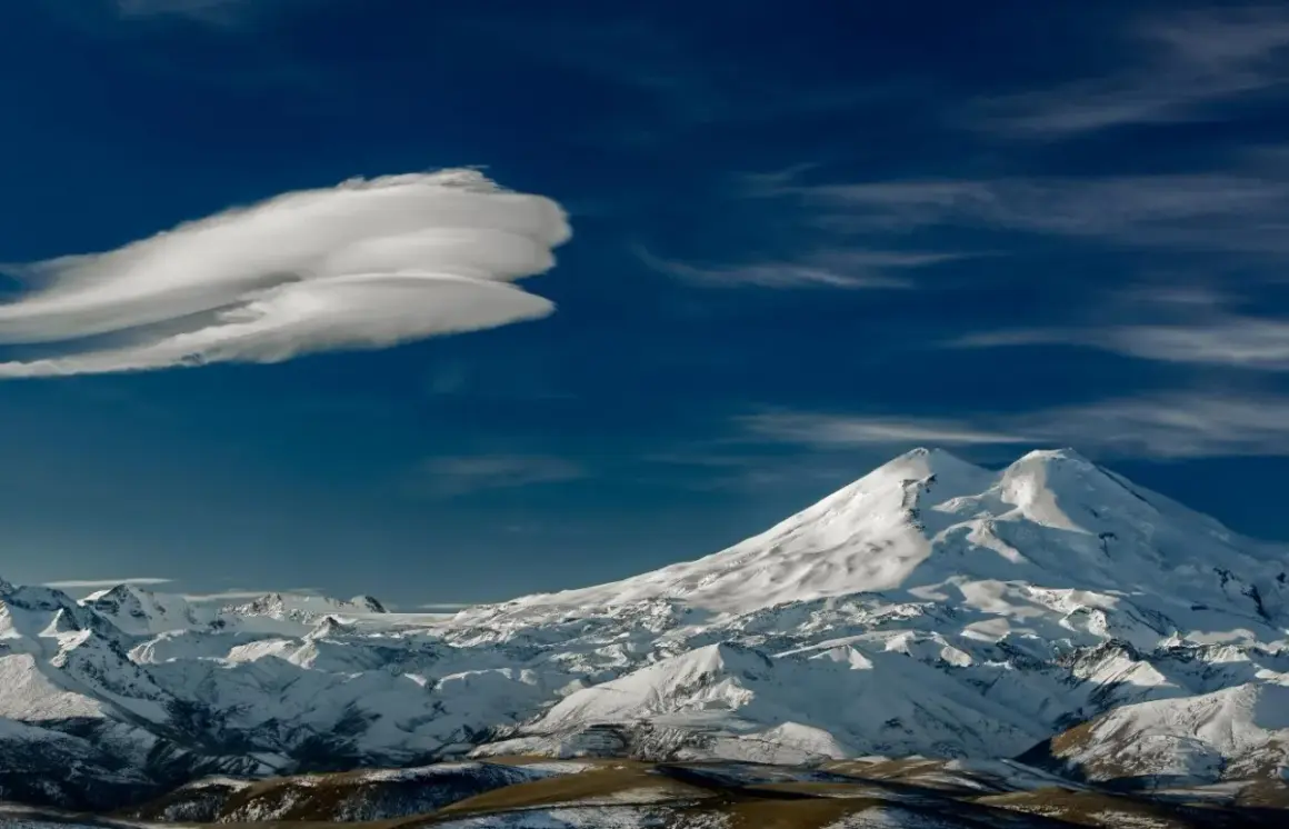 Pokryty śniegiem najwyższy szczyt Kaukazu, Elbrus, góruje nad ośnieżonymi pasmami górskimi pod błękitnym niebem z charakterystyczną chmurą.